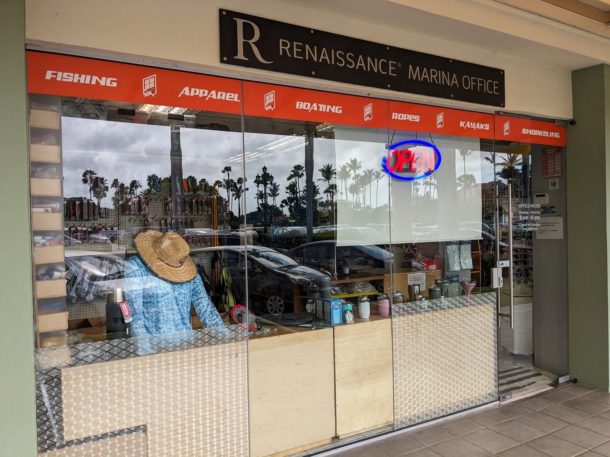 Exterior of a marina office with a storefront displaying a mannequin dressed in a blue shirt and straw hat, surrounded by various boating and fishing equipment. The glass window reflects palm trees and parked cars outside, while a neon 'Open' sign is visible above the door.