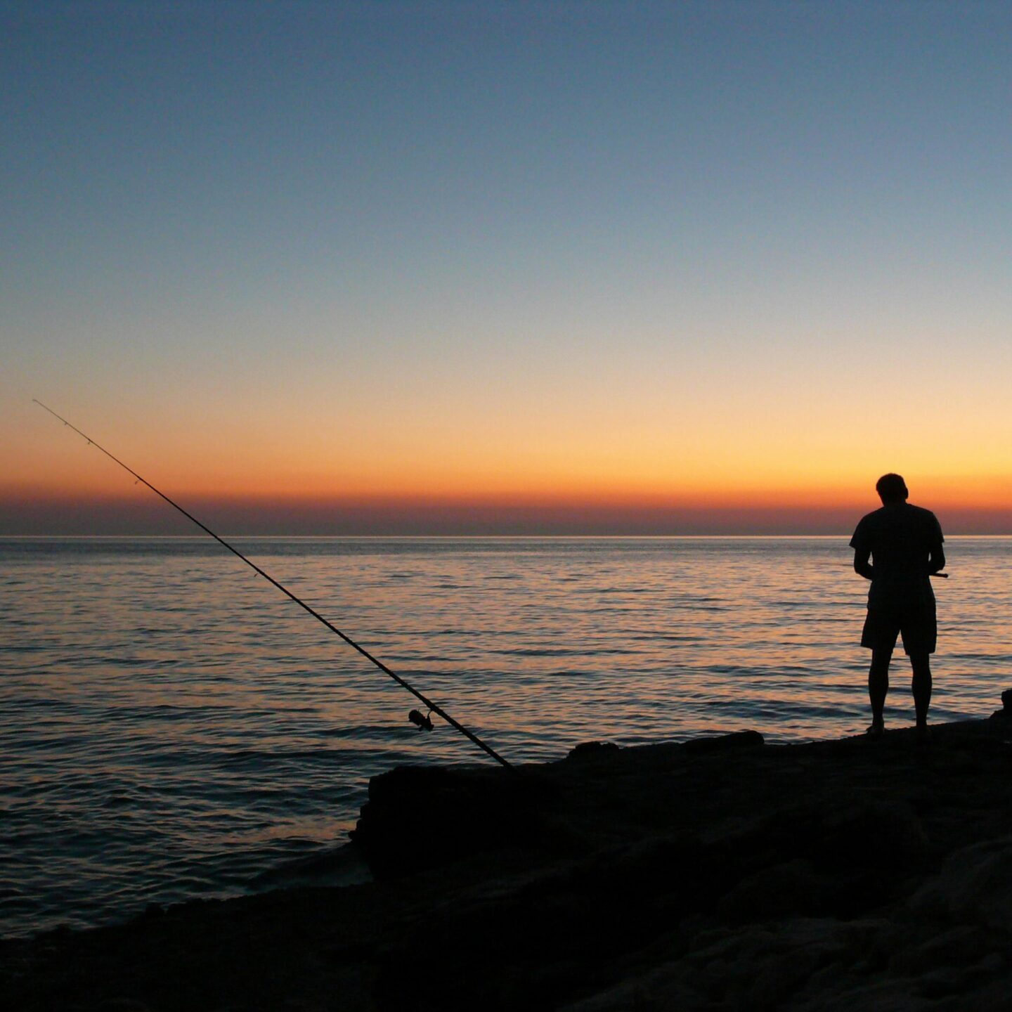 Silhouette of a person fishing at the edge of a calm sea during a vibrant sunset, with the sky transitioning from deep blue to warm orange hues.